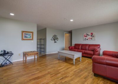 A living room with red leather sofas, a wooden coffee table, a small table with electronic devices, and artwork on light-colored walls. The floor is wooden, and a doorway leads to another room.