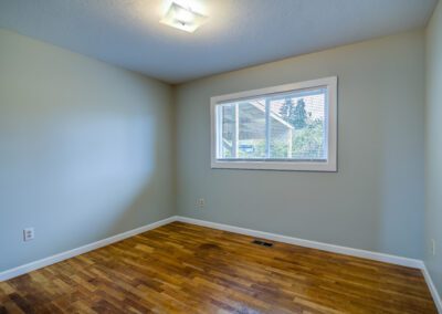 A vacant room with light-colored walls, a ceiling light, a window, and a hardwood floor.