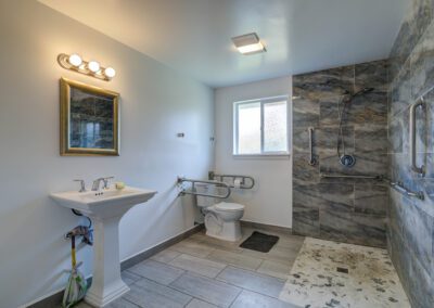 A modern accessible bathroom with a pedestal sink, a toilet with support bars, and a spacious walk-in shower area equipped with grab bars. The walls and floor feature grey marble tiles.