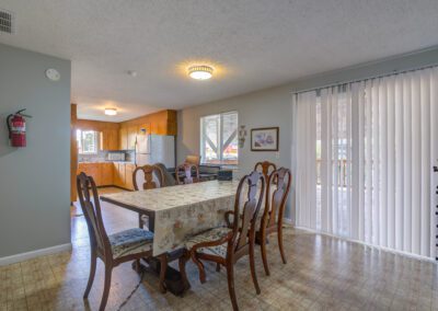 A dining area with a wooden table and six chairs, a floral tablecloth, and a view of the kitchen in the background. Vertical blinds cover the large window on the right.