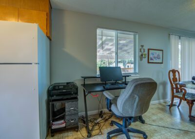 A small home office setup with a computer desk, monitor, chair, and printer in the corner of a room near a window. Next to it is a dining area with a wooden table and chairs, and a refrigerator.