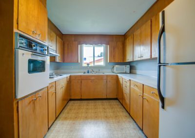 A kitchen with wooden cabinets, a white oven, a microwave on the counter, and a window above the sink. The floor is covered with patterned linoleum.