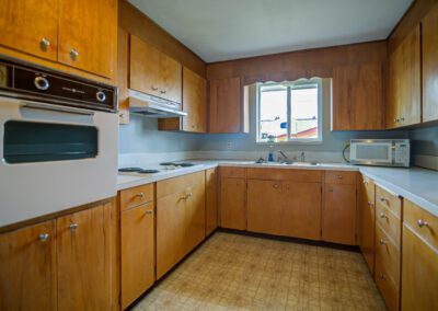 A kitchen with wooden cabinets, a white countertop, built-in oven, electric stovetop, microwave, sink, and a window. The floor has a patterned linoleum covering.