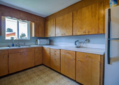 A kitchen with wooden cabinets, a microwave, kettle, and refrigerator, white countertops, and a window above the sink. The floor has a patterned design.