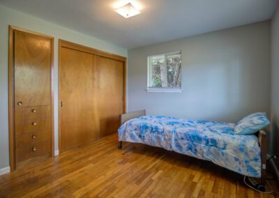 A small bedroom with wooden floors features a single bed with blue and white bedding, large wooden closet doors, a built-in wooden cabinet, and a small window.