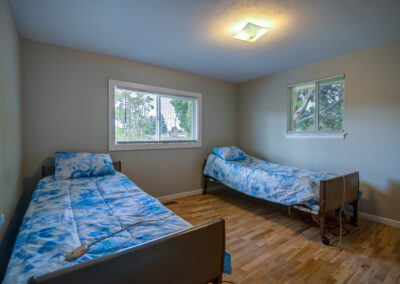 A small bedroom with two twin beds, each covered with blue and white bedspreads. The beds are positioned against adjacent walls under two windows, and the floor is made of light-colored wood.