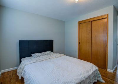 A modest bedroom with a bed against a light gray wall, white bedding, and wooden closet doors in the background.