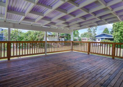 A covered wooden deck with a high roof and railing, overlooking a grassy backyard with trees and houses in the distance.