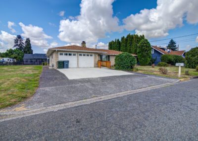 A one-story house with a two-car garage, two green trash bins, a concrete driveway, and a small front porch, on a clear day with clouds in the sky.